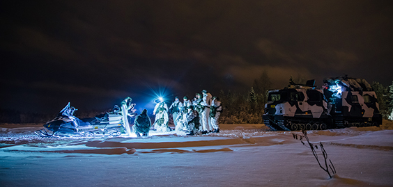 Sotilaita lumipuvuissa talvisessa mastossa moottorikelkan ympärillä. Vieressä tela-ajoneuvo.