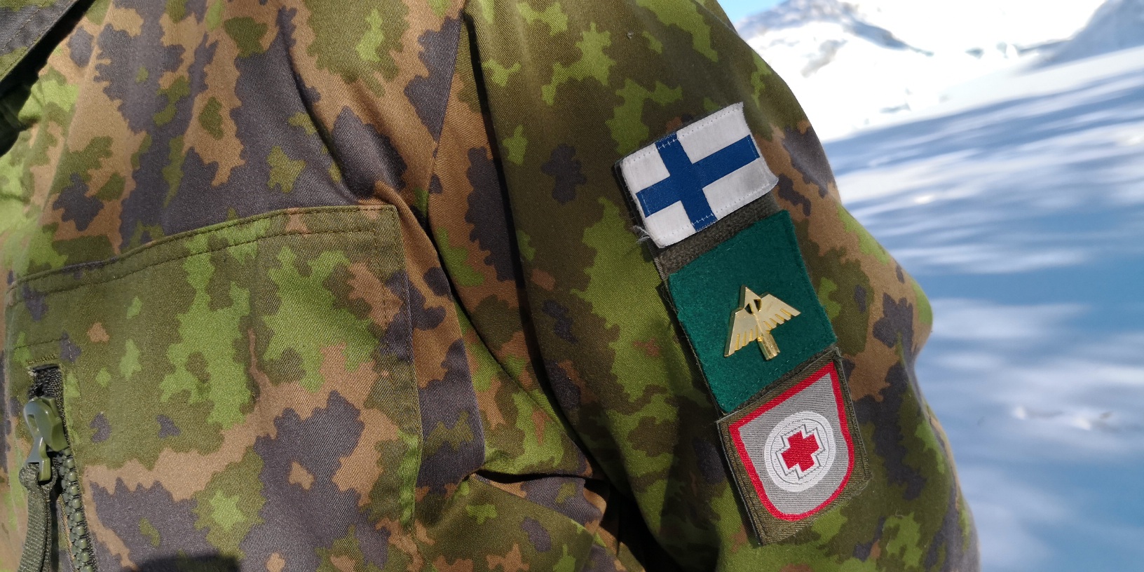 A soldier's suit sleeve with a Finnish flag, an arrow with wings and a red cross inside a white ball.