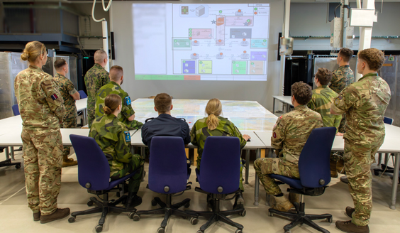 A group of people of different nationalities wearing camouflagr uniforms are looking at charts projected onto a large screen. Everyone has their back to the camera.