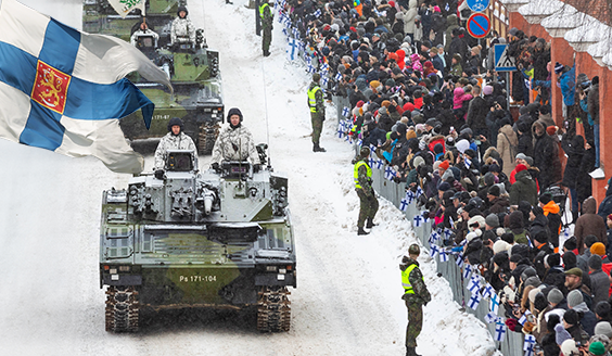 Kolme panssarivaunua ajaa peräkkäin lumisella tiellä. Vasemmassa kulmassa suomen lippu. Oikealla yleisöä katsomassa ohimarssia.