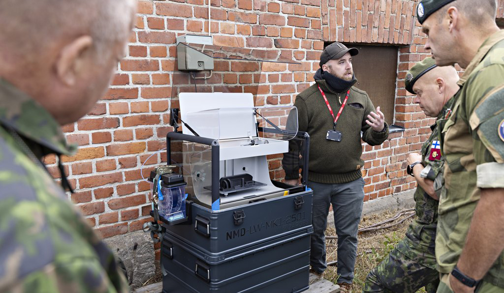 In the picture, four men – three soldiers and one civilian – are examining a 3D printer.