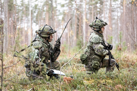 Kaksi aseistettua sotilasta Taipalsaaren harjoitusalueen maastossa, polviasennossa. toinen puhuu radiopuhelimeen.