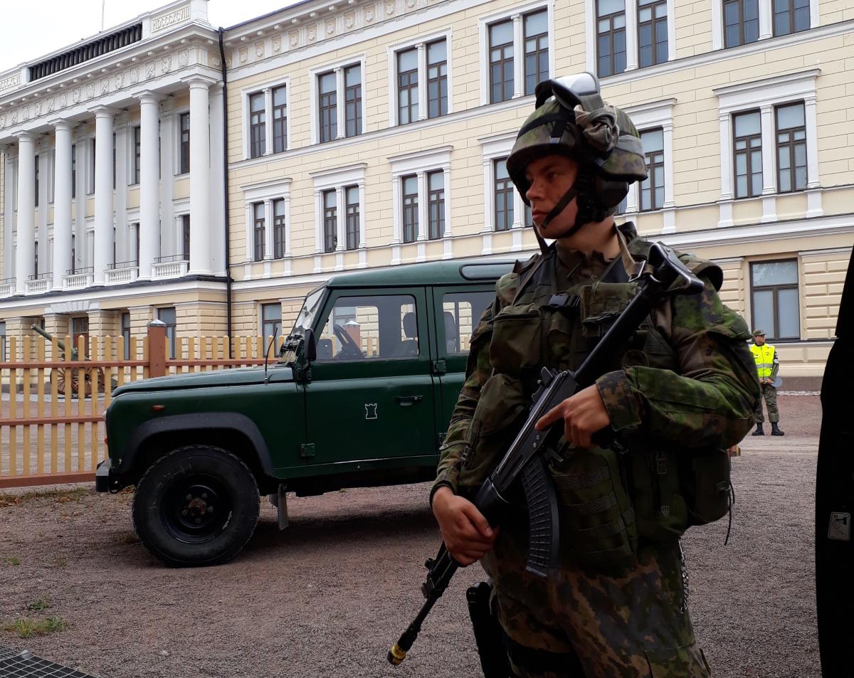 Maastopukuinen sotilas rynnäkkökiväärin kanssa vartioimassa Puolustusvoimien ajoneuvoja RUK:n päärakennuksen edessä.