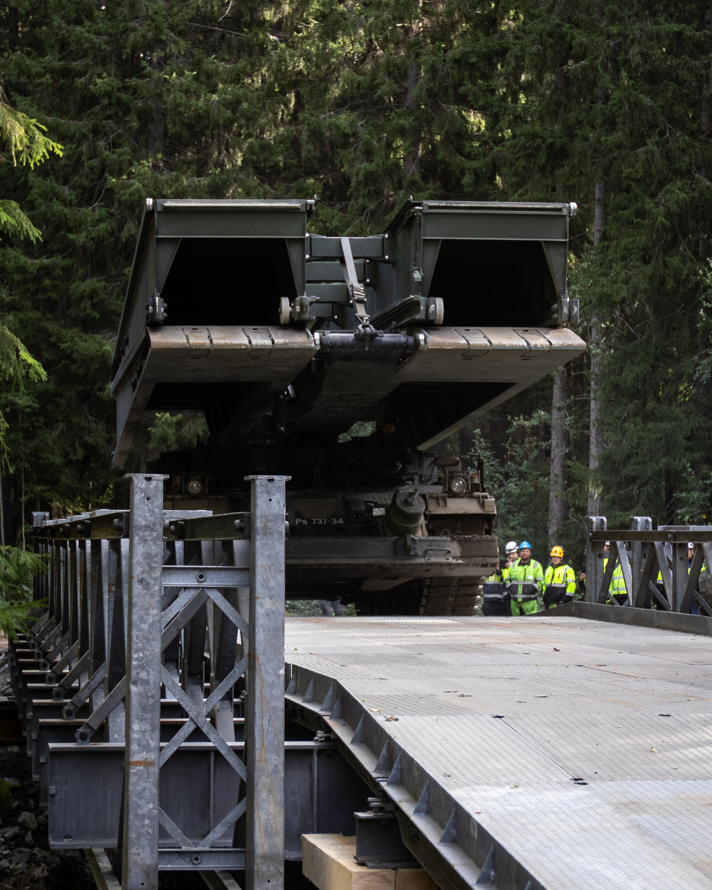 Siltapanssarivaunu ylittämässä palkkisiltaa, taustalla metsää ja huomiovaatteissa olevia ihmisiä