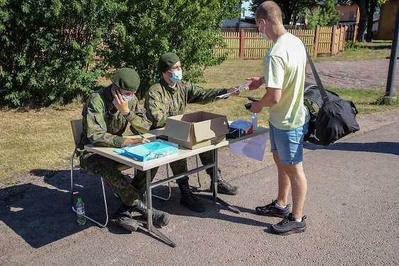 Maastopukuiset kaksi varusmiestä ottavat siviilivaatteissa ilmoittautuvan uuden alokkaan vastaan inttiin, hellettä pukkaa.