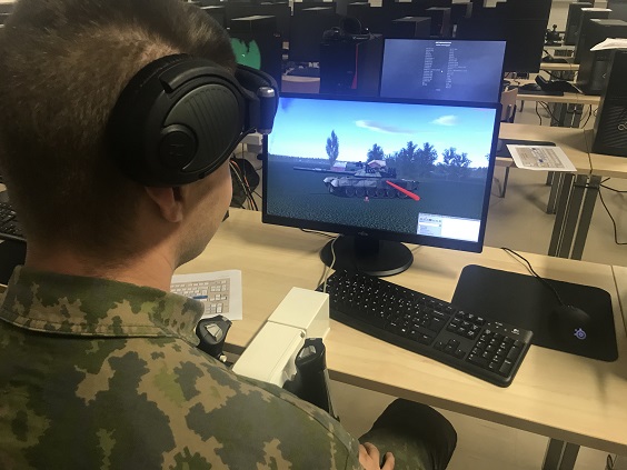 Sotilas käyttää panssarivaunusimulaattoria, maastopukuinen henkilö selin tietokoneen ääressä kuulokkeet korvilla, tietokoneruudulla panssarivaunu.