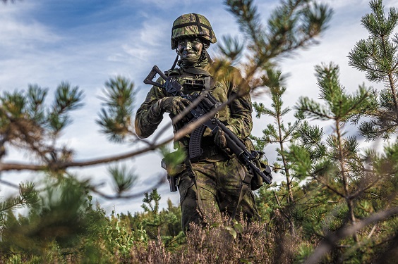 Maastopukuinen sotilas aseen kanssa polviasennossa kesäisessä maisemassa.
