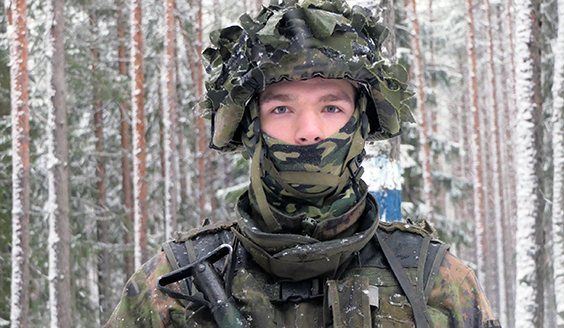 Lähikuva sotilaasta, taustalla luminen havumetsä