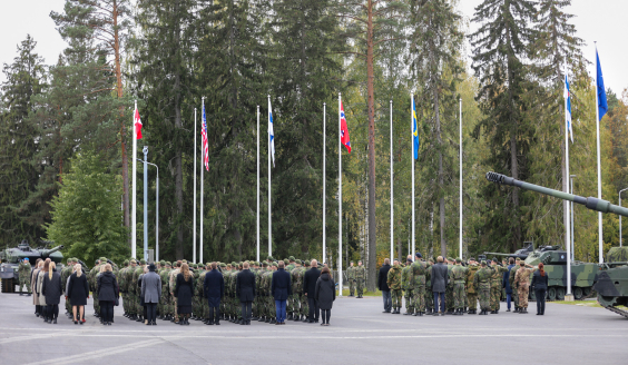 Staff standing with their backs to the camera.  The flags of Denmark, the United States, Finland, Norway, and Sweden fly behind the staff. The flags of Finland and NATO are also visible in the background on the right.