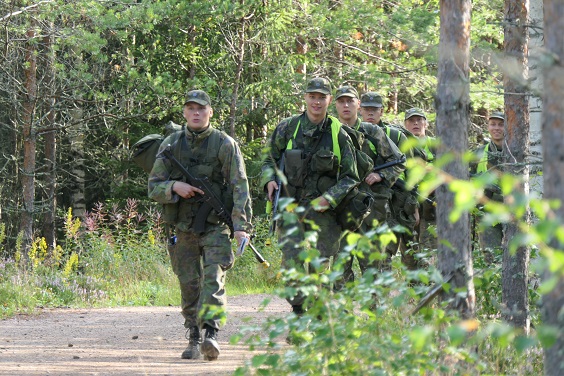 Varusmiehiä kävelemässä maastossa aseistettuna ja maastopuvuissa, hymyssä suin.