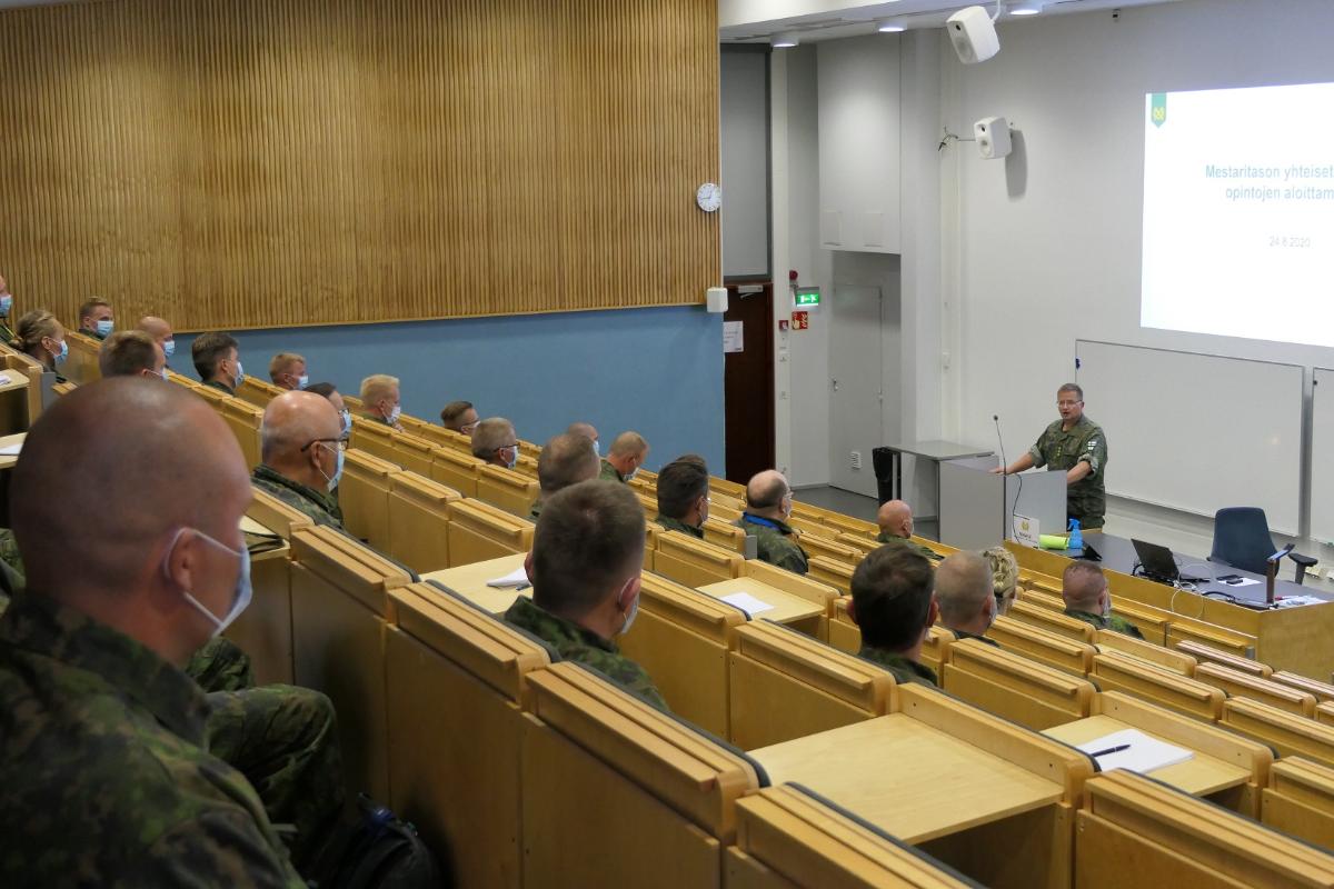 Salissa istuu turvavälit ja kasvomaskit muistaen sotilaita, jotka katsovat puhujaa, maastopukuista everstiä.