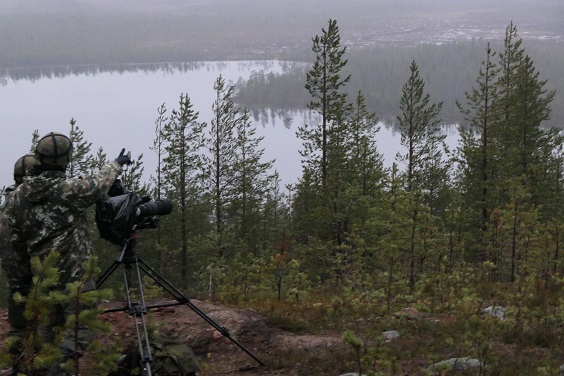 Tutkija maastopuvussa kameran kanssa tarkkailemassa ammuntoja Rovajärven maisemissa.