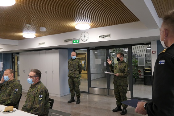 Sotilaita maastopuvuissa seisomassa sisätiloissa: kapteeni Laitinen kuuntelee, kun eversti Pietiläinen lukee perusteita Laitiselle myönnetystä sotilasansiomerkistä.