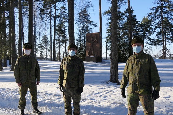 Kolme maastopukuista sotilasta seisoo lumisessa maisemassa, taustalla näkyy muistomerkki.