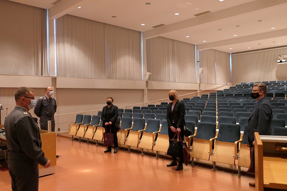 Isossa miltei tyhjässä auditoriossa seisoo turvavälien päässä toisistaa maskien kera kolme siviiliä, ja kaksi harmaapukuista sotilasta. Tutustumisvierailu Maasotakoulussa menossa.