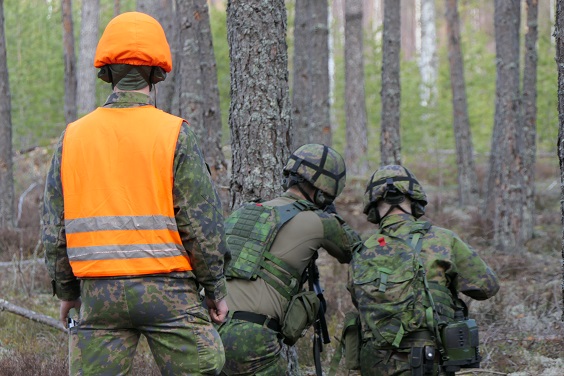 Maastossa sotilaita harjoituksessa, heidän takanaan kouluttaja oranssissa liivissä.