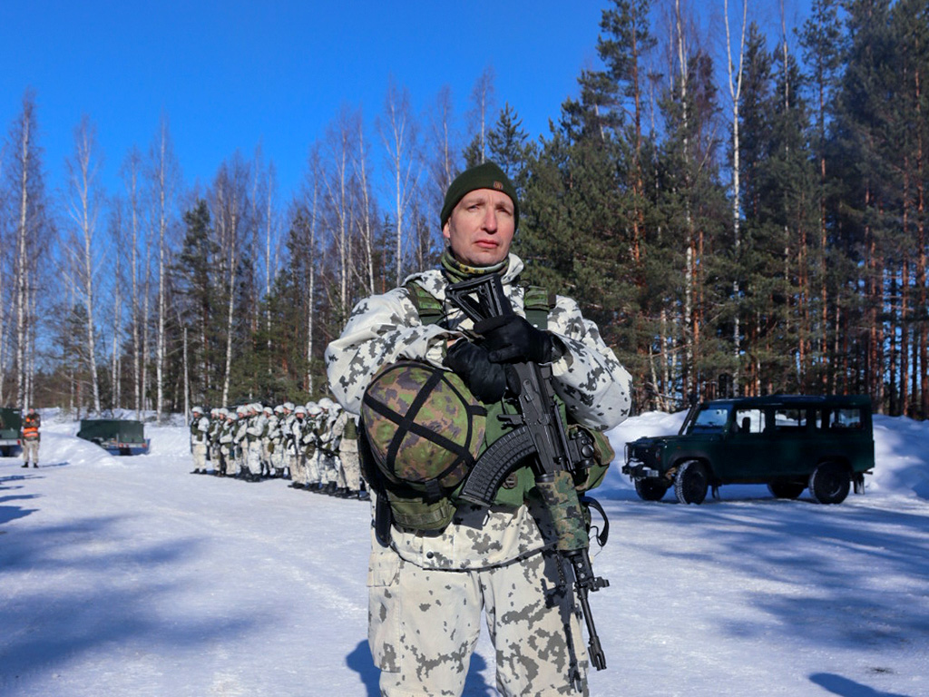 Talvipukuinen sotilas seisoo pidellen asettaan edessään, taustalla talvipukuisia taistelijoita rivissä talvisessa maisemassa