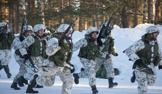 Maastopukuisia reserviläisiä aseistettuina juoksemassa lumisessa maastossa.