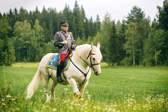 Musiikkimajuri Riku Huhtasalo rakuuna-asussa ratsastaa valkoisella hevosella kauniissa vehreässä maisemassa.