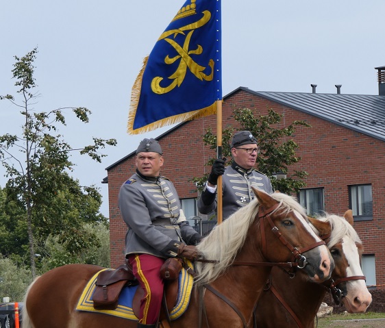 Kaksi rakuunapuvussa olevaa ratsastajaa hevosten selässä, rakuunalippu liehuu toisen kädessä.