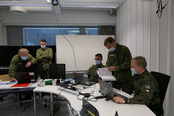 Sotilaita maastopuvussa esikuntatyössä tietokoneiden kanssa. Yksi seisoo toisen vieressä kirja kädessä. Muut istuvat tietokonepöytien äärellä.