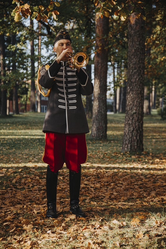 Sotilastrumpetisti soittamassa syksyisessä maisemassa, yllään rakuunoiden perinteisen tyylinen, harmaa luurankotakki ja punaiset hurmahousut.