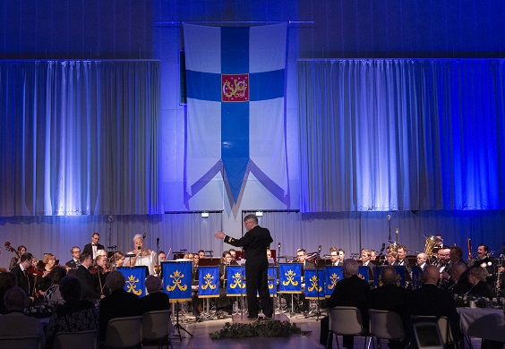 Iso Puolustuvoimain lippu seinällä, orkesteri soittaa ja kapellimestari johtaa.
