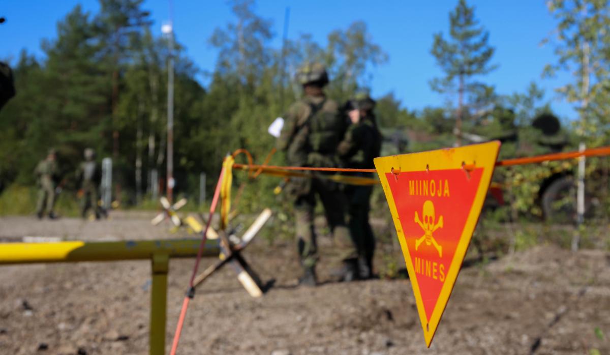 Miinoista varoittava lippu roikkuu keltaisella langalla, taustalla kaksi sotilasta.