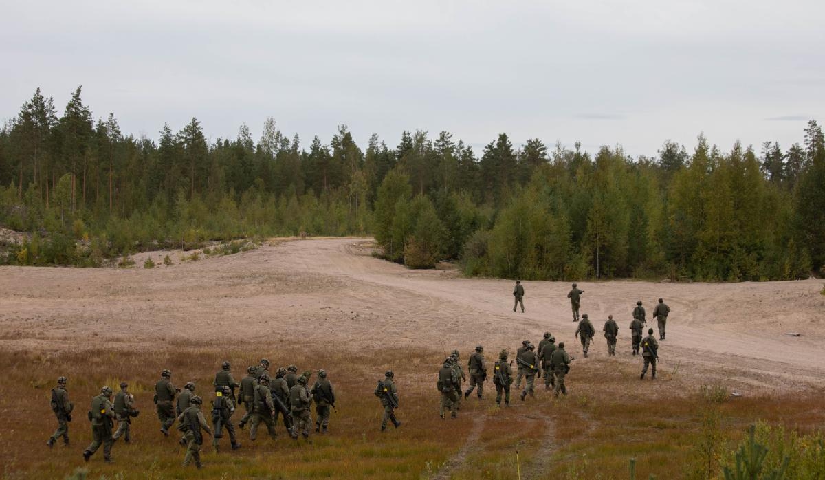 Iso joukko sotilaita kävelee hiekkakentällä kantaen aseitaan, taustalla metsää.