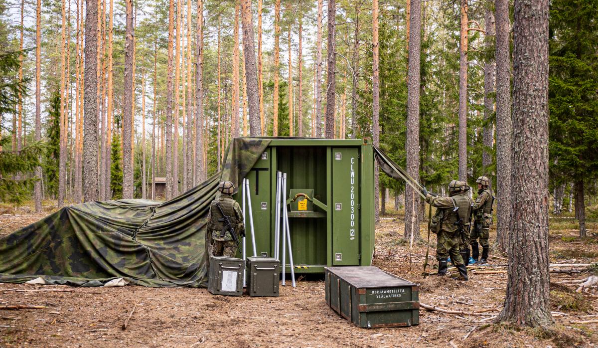 Vihreä merikontti metsässä, sotilaat vetävät naamioverkkoa kontin päälle.