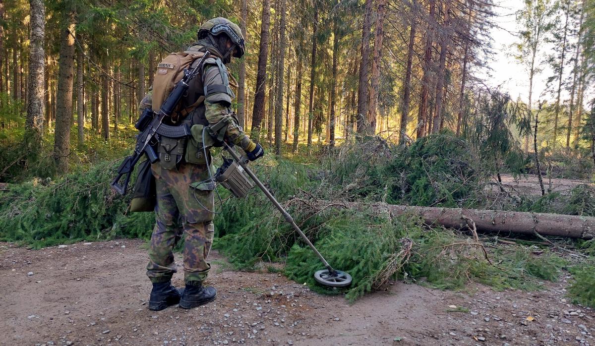 Sotilas metsätiellä, miinaharava käsissään haravoi maata tielle kaadettujen puiden luota