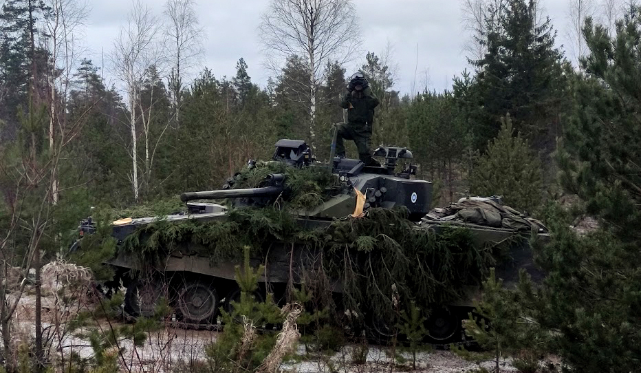 Sotilas seisoo panssarivaunun päällä ja kiikaroi kohti kameraa, taustalla metsää.
