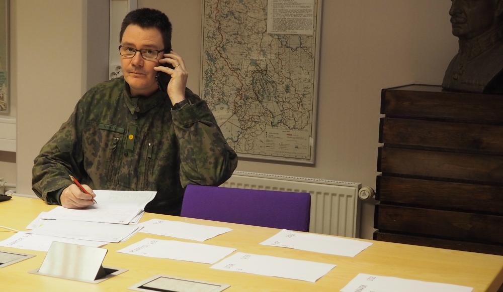 Sotilaspukuinen mies puhuu puhelimeen ja katsoo kameraa kohti. Taustalla kartta ja pöydällä papereita.