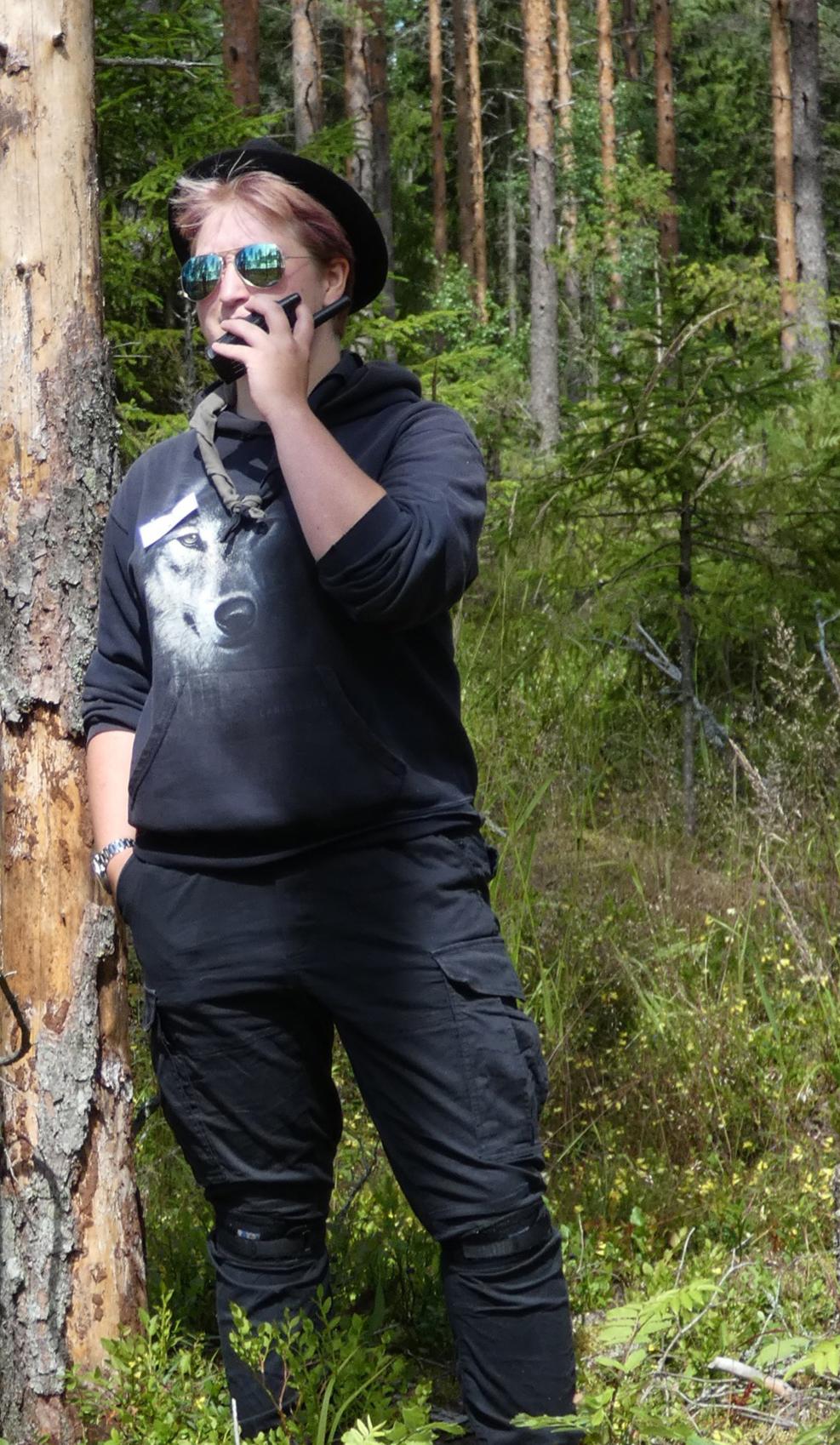 tummiin pukeutunut nainen nojaa puuhun ja puhuu radiopuhelimeen, taustalla metsää.