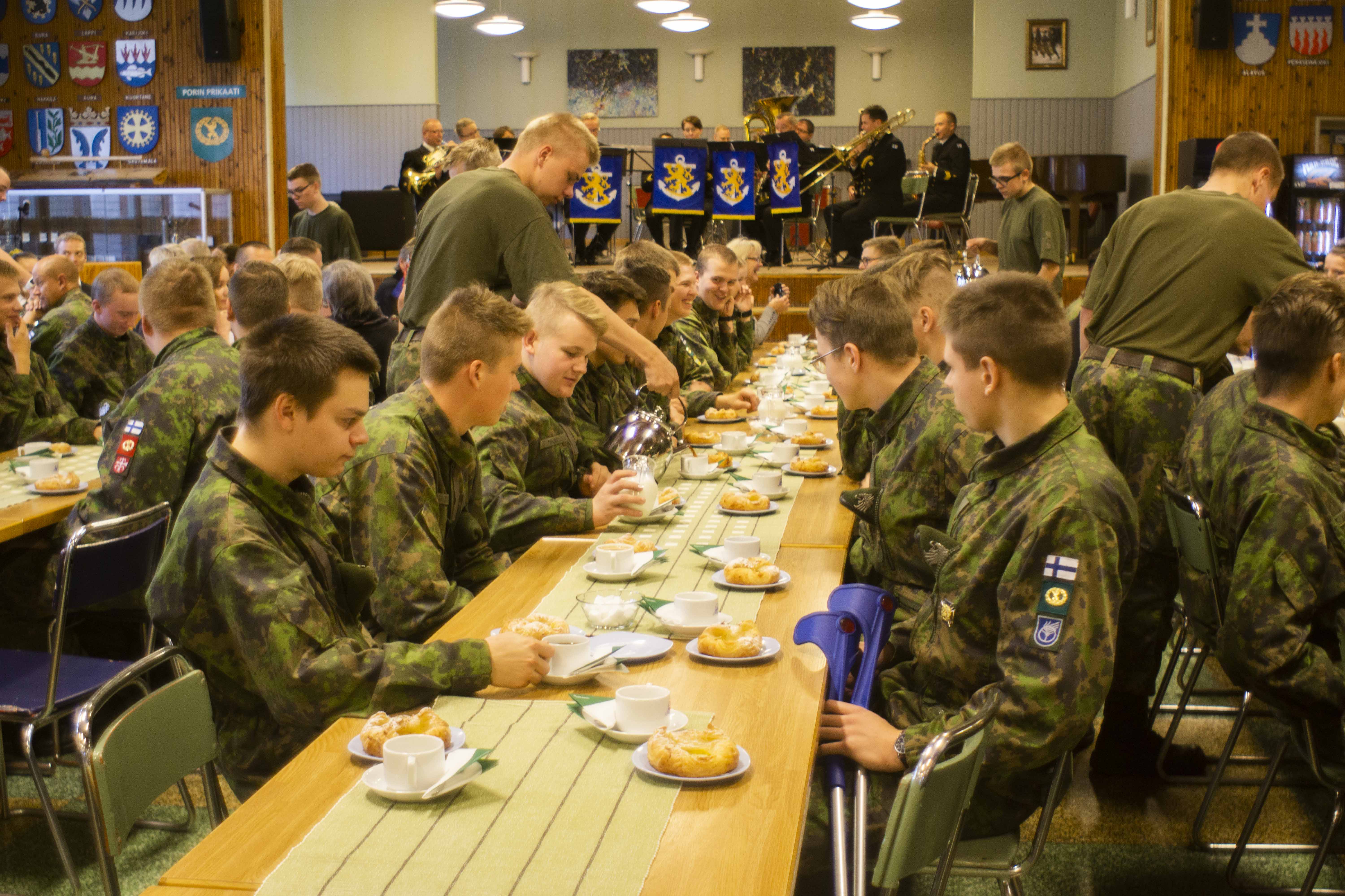 Sotilaat syövät pullaa ja juovat kahvia sotilaskodissa, taustalla soittokunta soittaa.