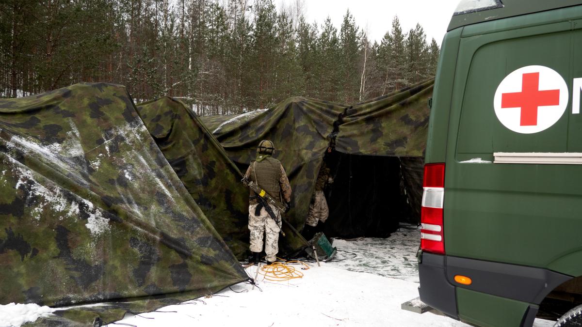 vasemmassa kulmassa edessä näkyy ensiapuauton perä ja punainen risti, taustalla pitkä teltta, sotilas seisoo edessä.