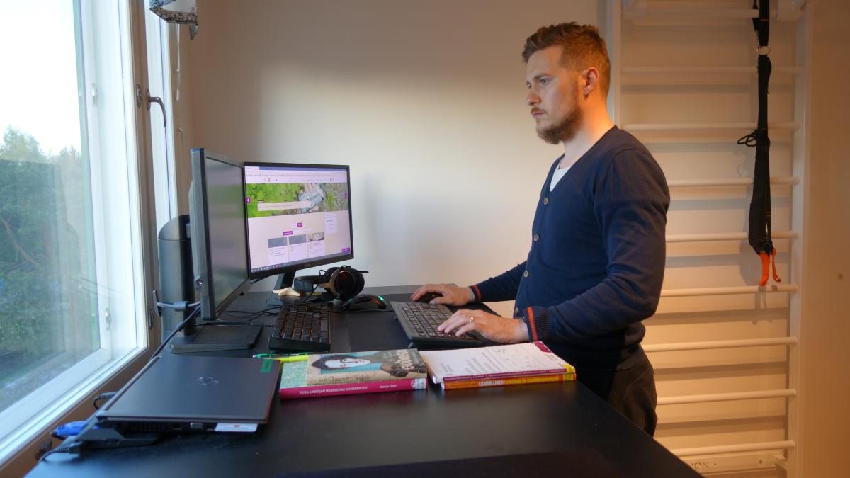 Tom Sandberg seisoo työpöydän äärellä, pöydällä tietokone, kirjoja ja vehreä maisema ikkunassa.