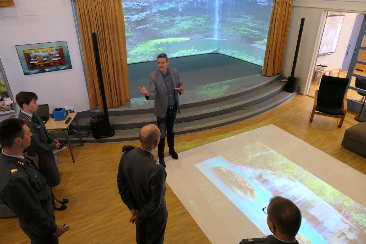Kuvassa Maasotakoulun delegaatio tutustuu rehtori Leppäsen esittelemään 360-ympäristöön eli luokkatilaan jossa seiniin heijastuu interaktiivista liikkuvaa kuvaa ja älylattiaan eli lattiaan heijastuvaan interaktiiviseen ruutuun.