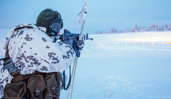 Sotilas tähtää rynnäkkökiväärillä maalitauluun lumisessa maisemassa, kädessään hänellä on myös hiihtosauvat