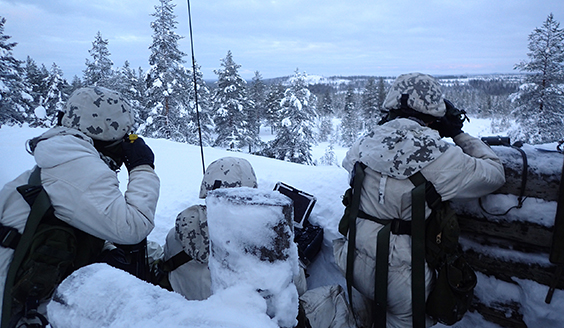 Kolme sotilasta poterossa lumisessa metsässä. Yksi tähystää kiikareilla, yksi käyttää tietokonetta ja yksi puhuu radiopuhelimeen.