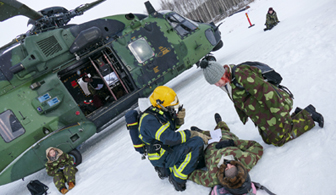 Sotilas makaa maassa ja palomies ja toinen sotilas ovat hänen vierellään kyykyssä. Taustalla helikopteri.