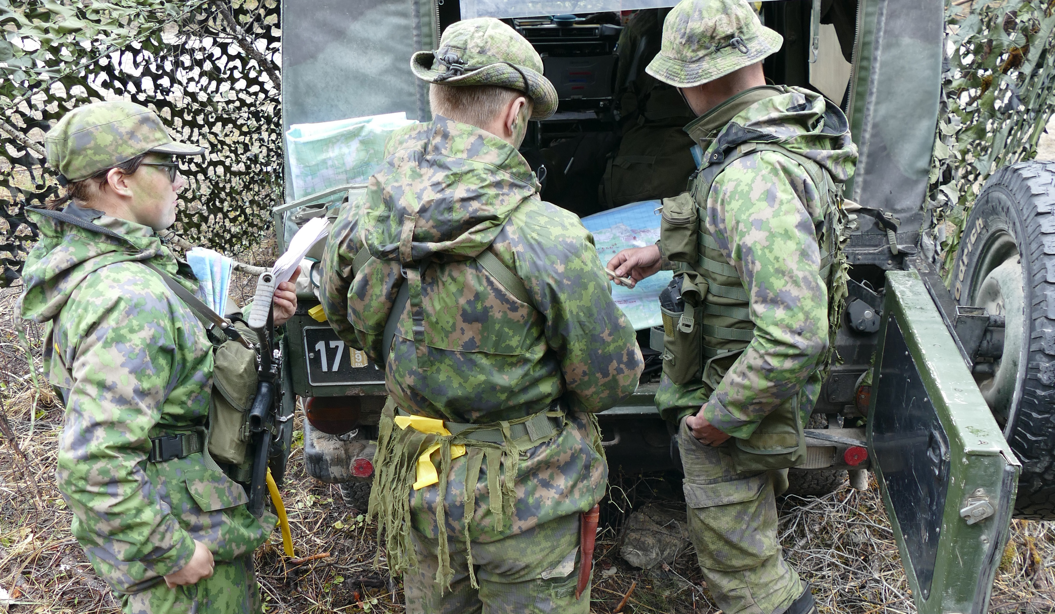 Sotilaat tutkivat karttaa maastoauton perässä, taustalla naamioverkkoa.