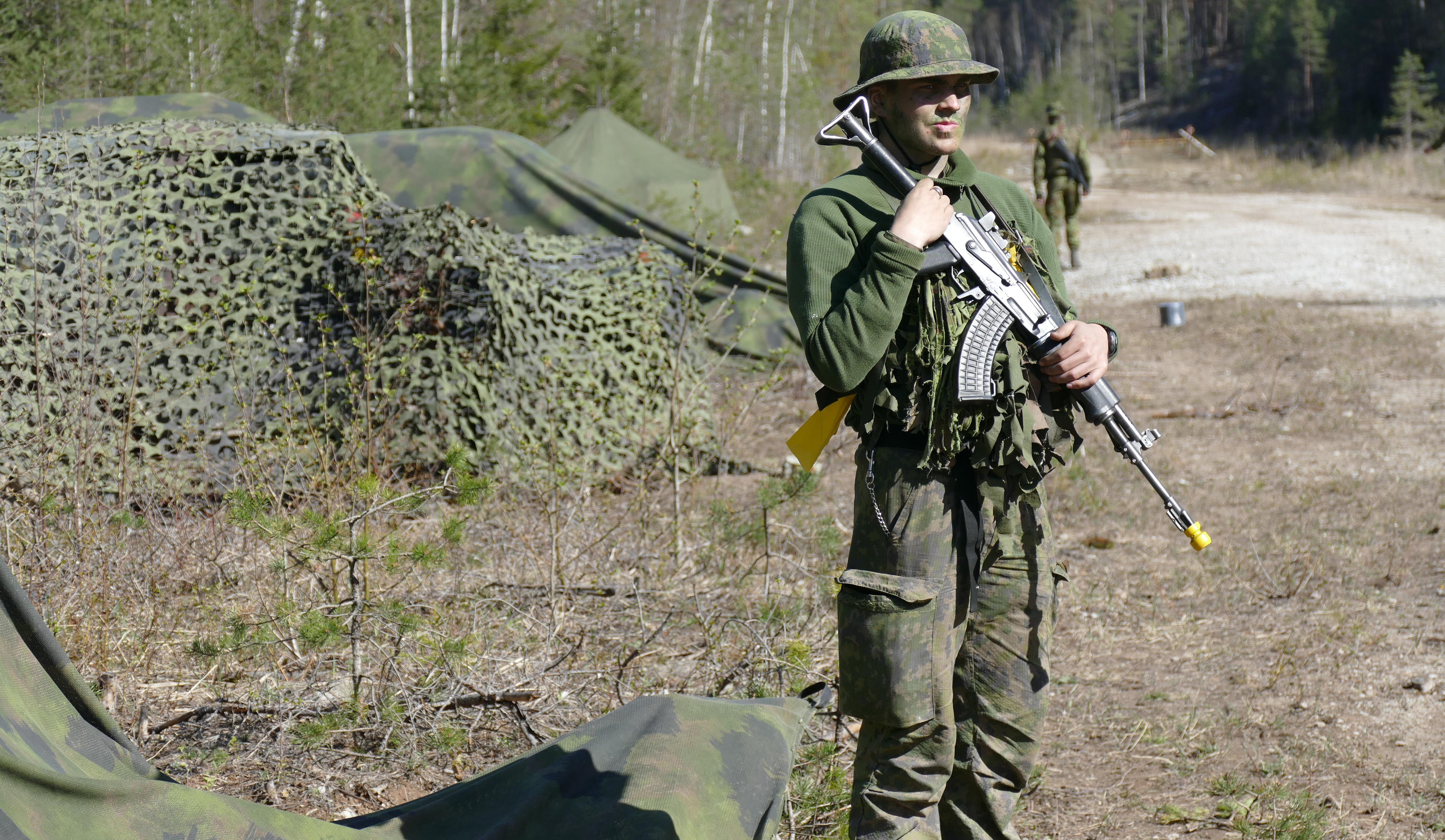 Sotilas seisoo rynnäkkökivääri rinnallaan auringossa, taustalla naamioitua kalustoa.