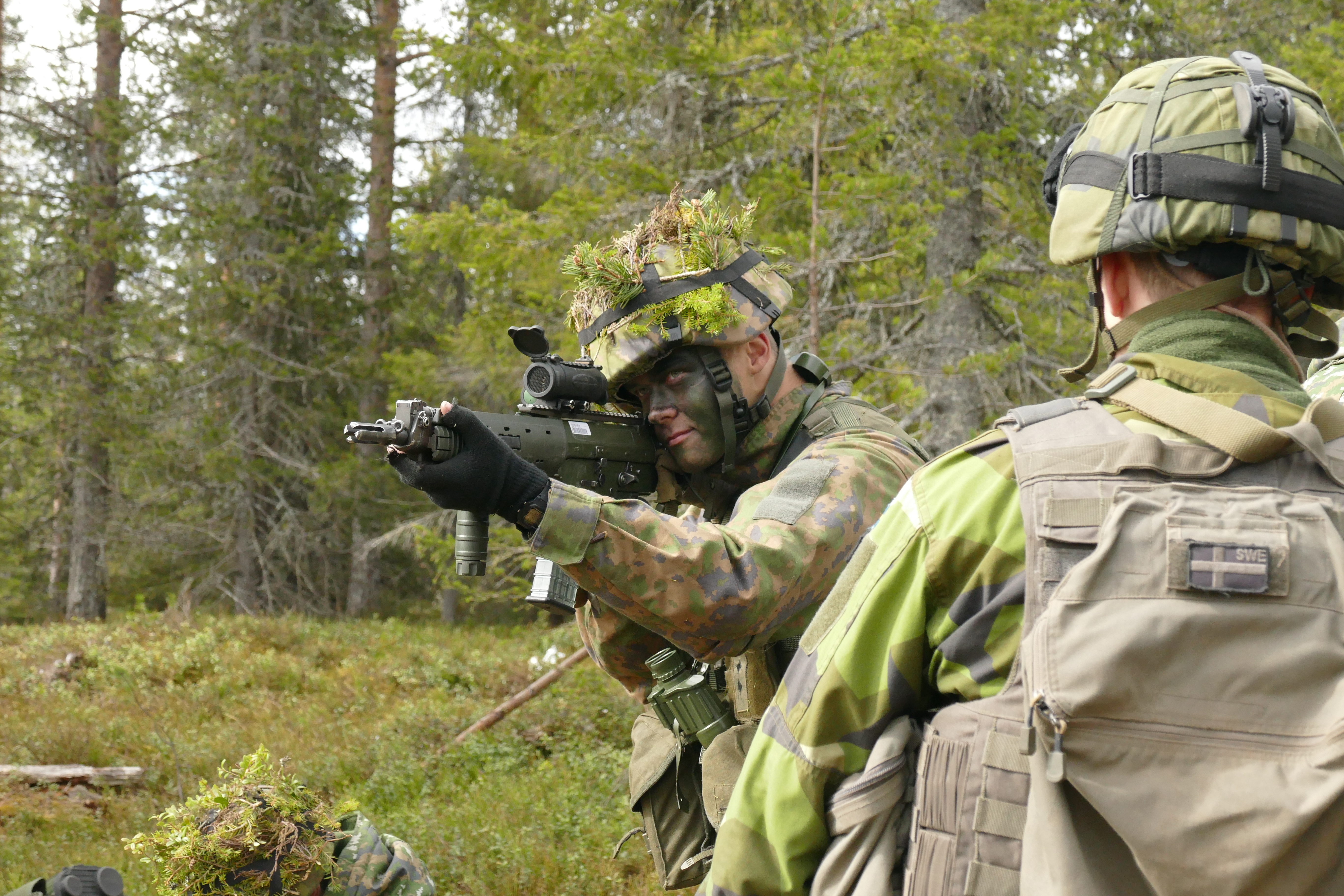 Suomalaisvarusmies tähtää ruotsalaissotilaan aseella, ruotsalaissotilas on selin.