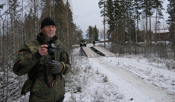 Saksalainen sotilashenkilö kamerakädessä ja taustalla suomalaiset sotilaat ylittämässä jokea juuri perustamansa sillan avulla.