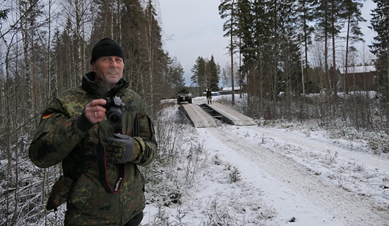 Saksalainen sotilashenkilö kamera kädessä ja taustalla suomalaiset sotilaat ylittämässä jokea juuri perustamansa sillan avulla.