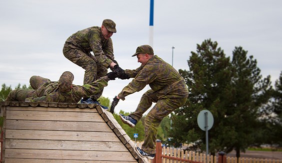 Kuvassa sotilas kiipeää esteradalla ylös ja toiset auttavat vetämällä häntä kädestä.