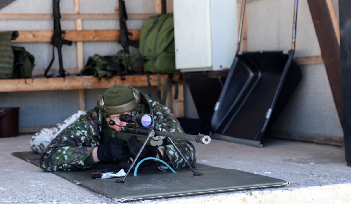 Miessotilas makaa vatsallaan betonilla ja tähtäilee tarkkuuskiväärin tähtäimen läpi, piippu osoittaa vähän ohi kamerasta.