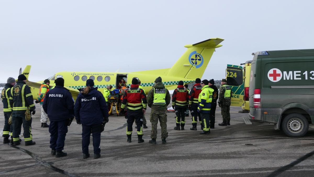 Potilasta nostetaan paareilla lentokoneeseen. Usean eri viranomaisen edustajat seisovat etualalla selin.
