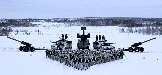 Kanoner, stridsvagnar och ett missilfordon symmetriskt sida vid sida, framför dem en stor grupp soldater i en triangulär formation. Snöig skoglandskap i bakgrunden.
